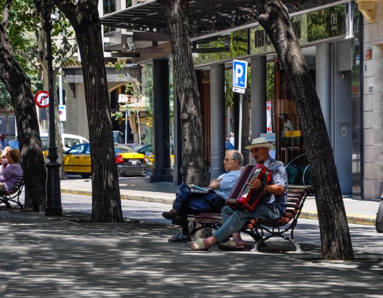 Barcelona Street Music; Photo: SPFischer Under the shade of trees, two men sit on a bench, one playing an accordion, the other relaxing and listening.