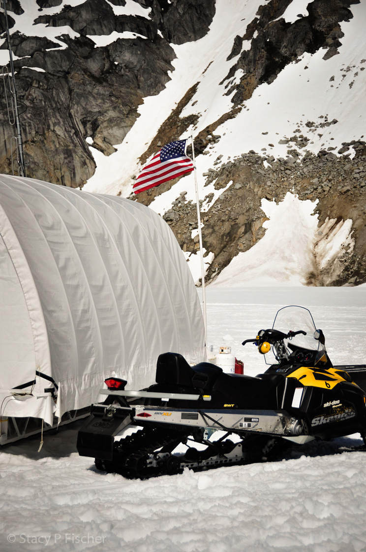 Waving in the breeze, an American flag atop a pole is the backdrop to a musher's tent and yellow snowmobile.
