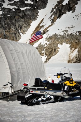 American Flag; Photo: SPFischer Waving in the breeze, an American flag atop a pole is the backdrop to a musher's tent and yellow snowmobile.