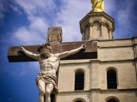 Against a deep blue sky, a gilded statue of Mary atop the Cathedral Notre Dame des Doms reaches downward toward a crucified Jesus.