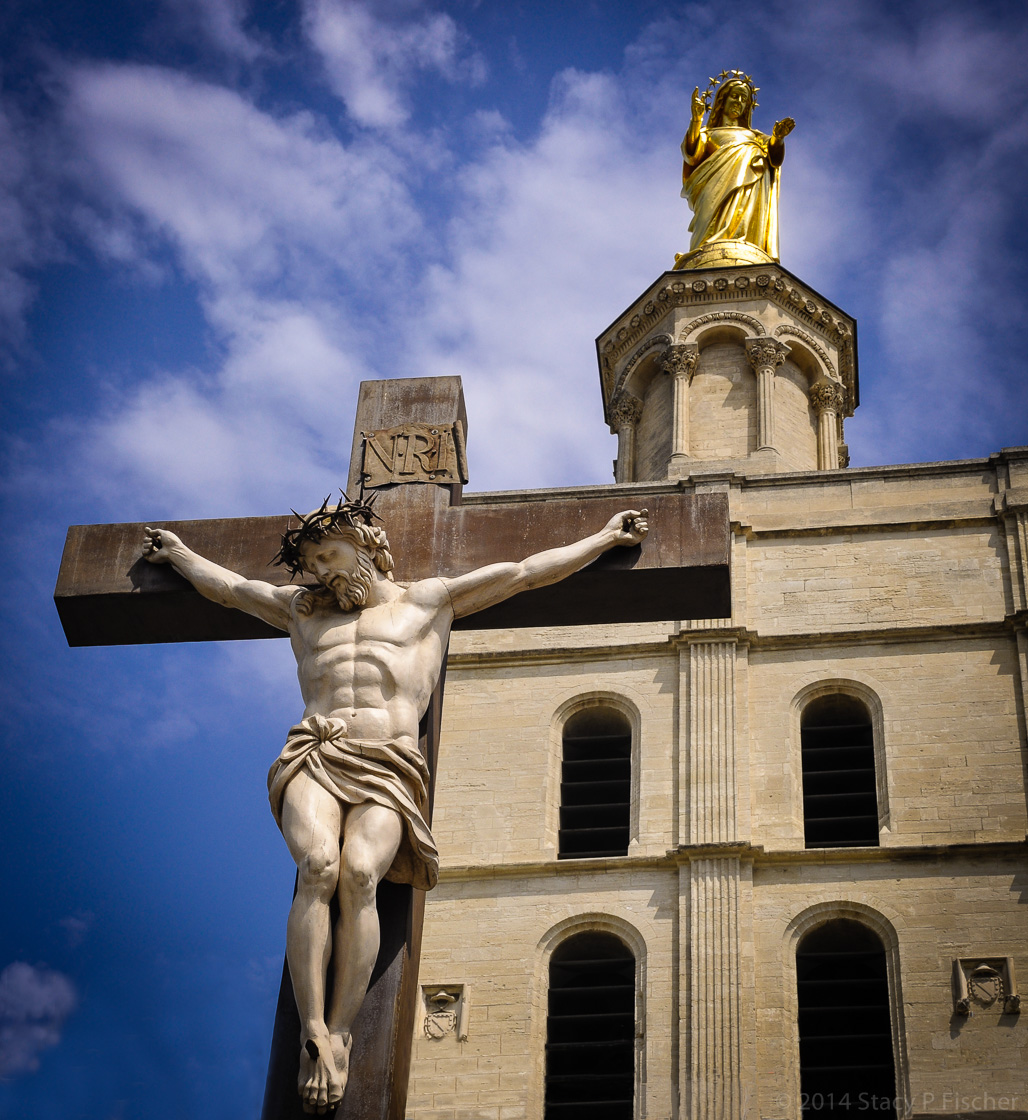 Against a deep blue sky, a gilded statue of Mary atop the Cathedral Notre Dame des Doms reaches downward toward a crucified Jesus.