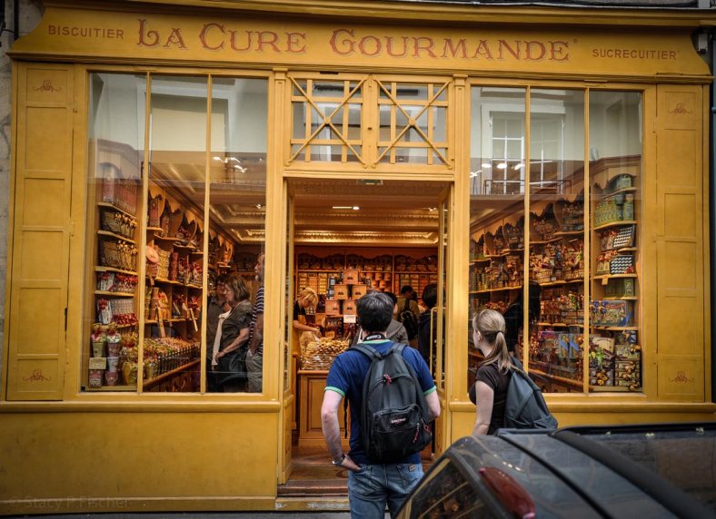 A couple stands on the sidewalk outside the door to a sweets shop, looking in, as if deciding whether or not to enter.