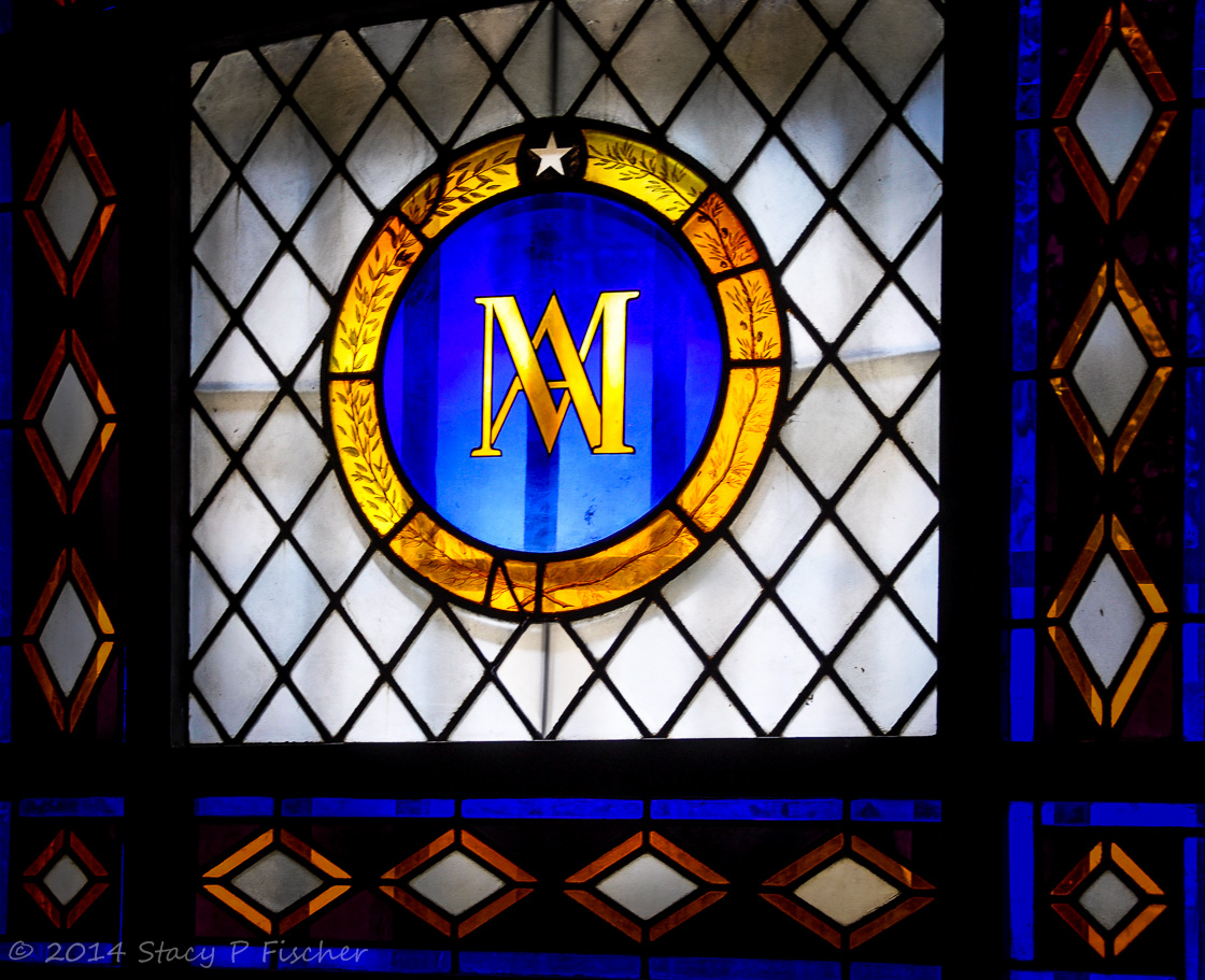 Marie Antoinette's gold initials in a deep blue circle, ringed by an outer gold circle containing olive branches, form a crest on a colorful window.