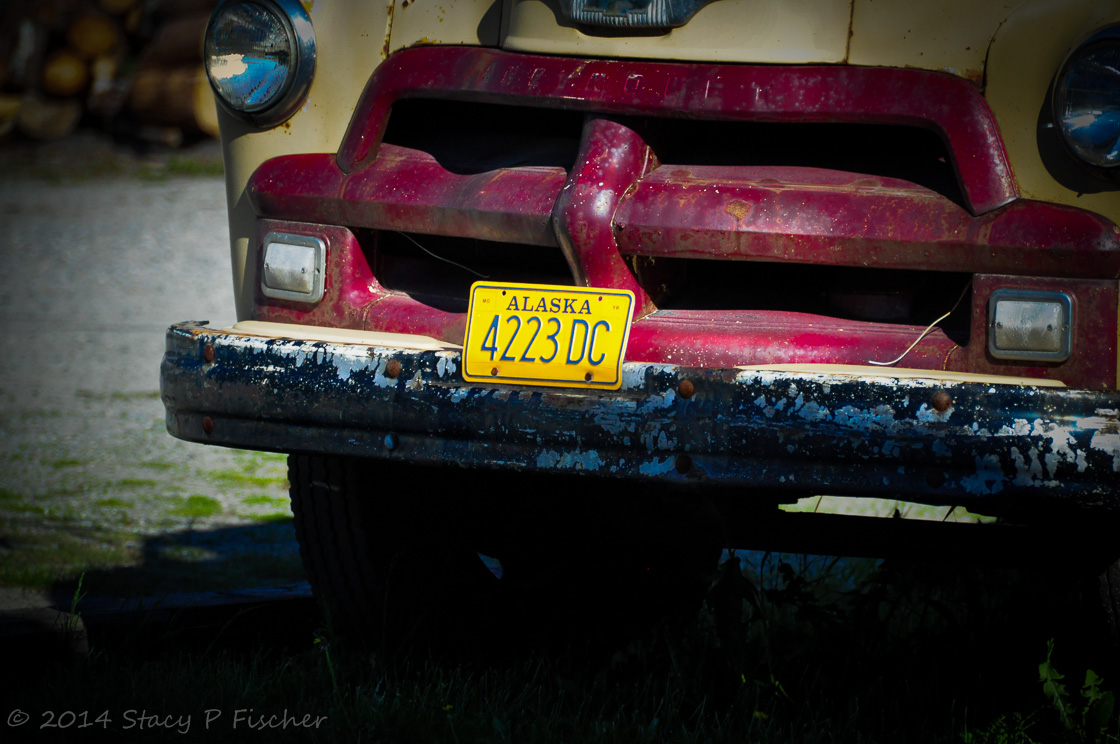 The grill of an abandoned red and beige vintage car still displaying an old gold Alaska license plate.