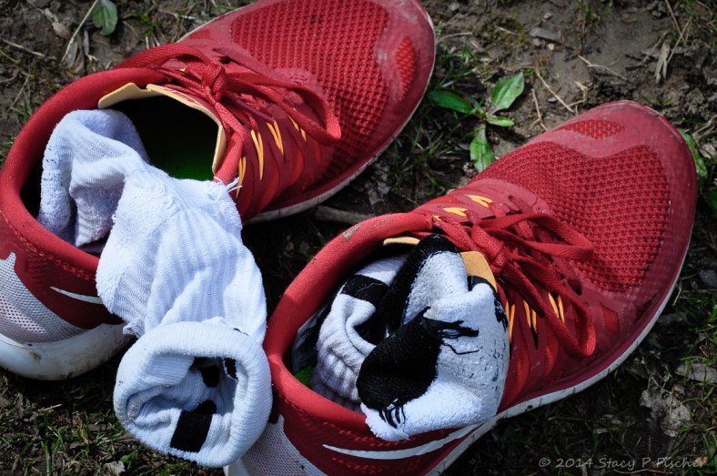 A pair of muddy red tennis shoes stuffed with white socks, sitting on wet ground with hints of green grass and weeds.