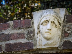 White stone relief of a woman's face and hair in the middle of a brick arch, green-leaved trees softly focused in the background.