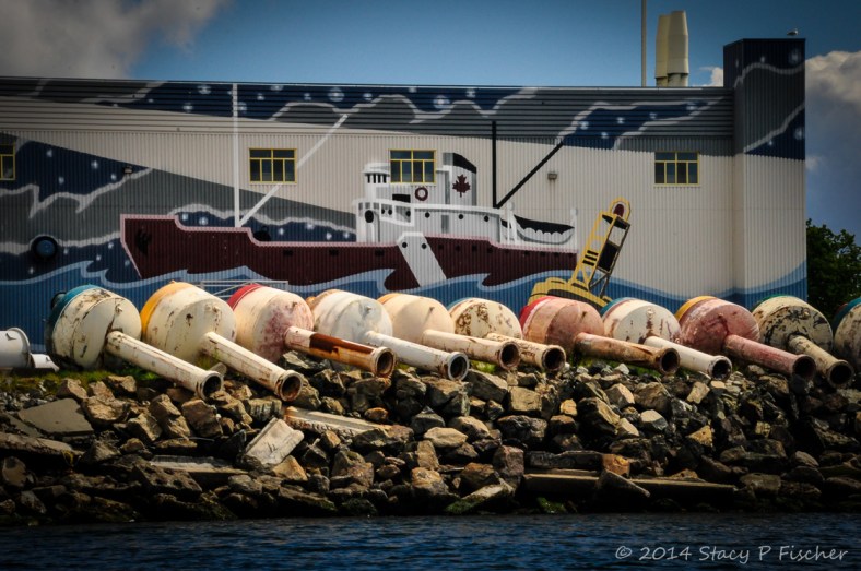 A row of ten weathered buoys on their sides in front of a mural of a tugboat on a waterfront building.