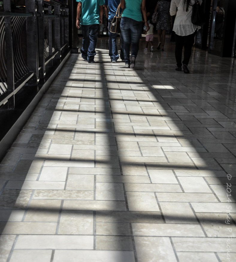 Square shadows fall in a vertical line on the floor of a mall with shoppers at the far end. 