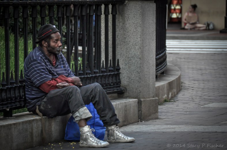 A forlorn-looking man in ill-kept clothing sits on a cement stoop, while across the street, a young woman holds a sign asking for help.
