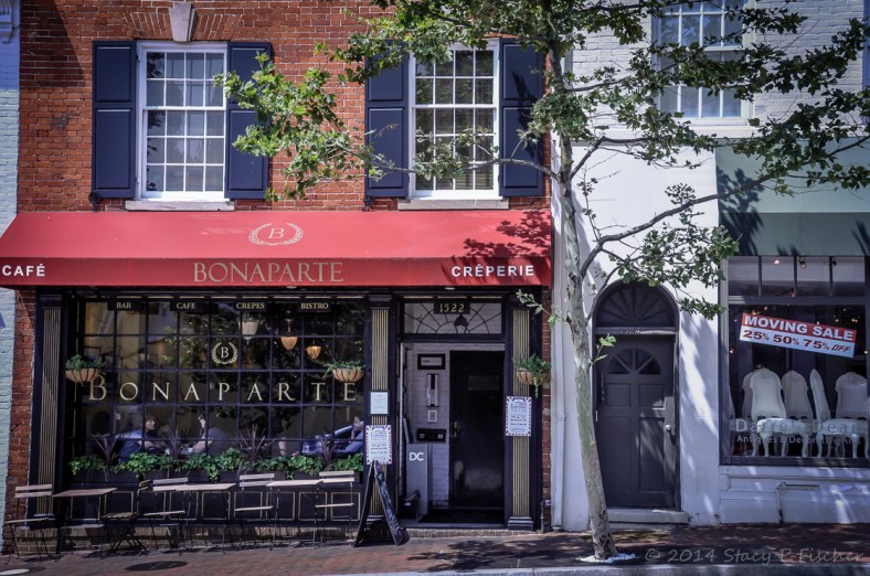 A red-awning hangs on the red-bricked facade of a French cafe, while its neighboring white-bricked furniture store advertises a moving sale.