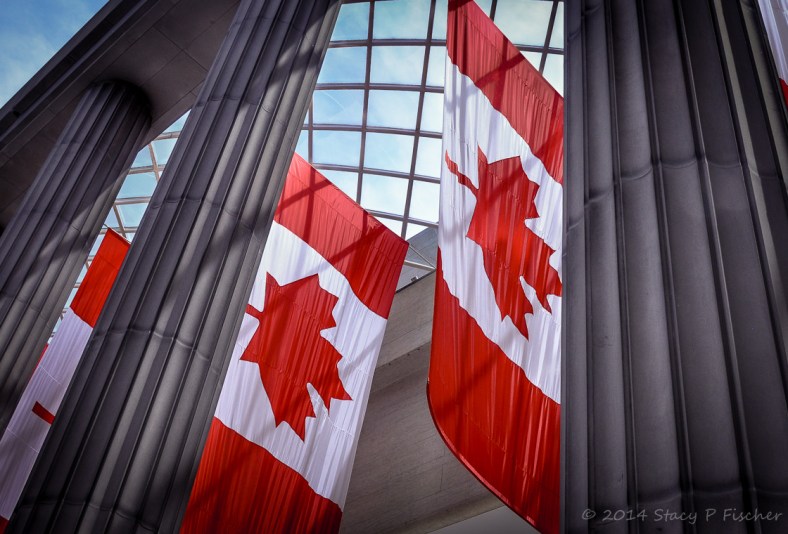 Canadian flags suspended horizontally, framed by marble pillars and overhead glass grid showing a blue sky.
