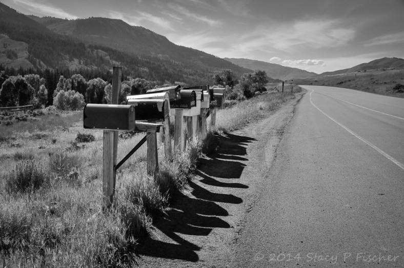 A row of mismatched rural mailboxes on posts and their shadows alongside a long two-lane road that winds into mountains in the distance.
