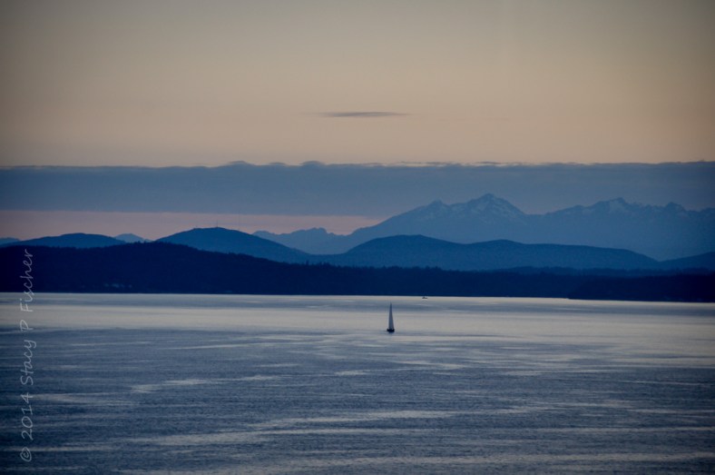 Dusk in Puget Sound creates layers of light and deep blue out of the sky, mountains, water, and lone sailboat. 