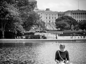 Woman sitting on the edge of a pond, reading, with US Capitol in the background.