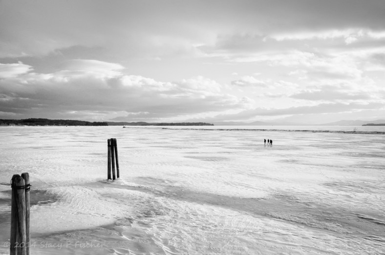 Lake Champlain, frozen over, with a group of three people in the distance walking on it.