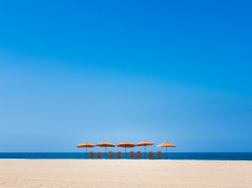 Orange umbrellas and chairs on a beach silhouetted against the bright blue of the sky and water.